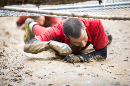 Teilnehmer am Wildsau Dirt Run mit rutschfesten uvex Schutzhandschuhen kriecht unter einem Netz durch den Schlamm