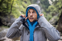 Mann mit warmer Outdoorjacke setzt sich die Kapuze seiner darüber getragenen, wasserabweisenden Funktionsjacke auf.