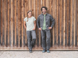 Mann und Frau mit nachhaltiger Arbeitskleidung vor Holzwand