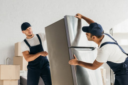 Zwei Möbelpacker in Latzhosen und Schirmmützen transportieren einen Kühlschrank.