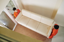 Zwei Umzugshelfer tragen eine cremefarbene Couch durch einen Flur.