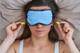Frau mit Schlafmaske liegt im Bett und steckt sich Gehörschutzstöpsel in die Ohren.