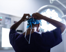 Chirurg steht im OP-Saal und befestigt die Haltebänder seiner Atemschutzmaske am Kopf.
