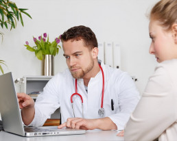 Arzt sitzt mit Patientin im Sprechzimmer und zeigt ihr etwas auf einem Laptop.