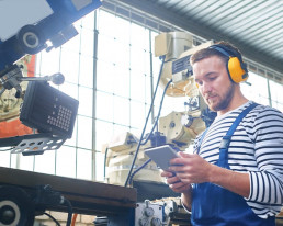 Arbeiter mit Arbeitskleidung und Gehörschutz steht in einer Industriehalle und blickt auf das Display eines Tablets.