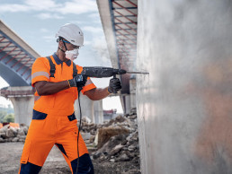 Bauarbeiter in uvex Schutzkleidung und Schutzausrüstung bohrt mit einer Schlagbohrmaschine ein Loch in eine Wand.