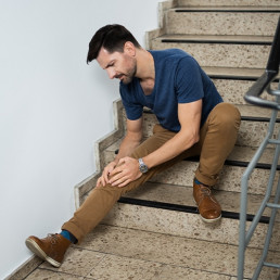 Mann sitzt in einem Treppenhaus auf den Stufen, streckt das rechte Bein von sich und hält sich das Knie.