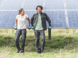 Frau und Mann in nachhaltiger uvex Workwear und mit nachhaltigen uvex Schutzhandschuhen lehnen an Solarpanelen auf einem Feld.