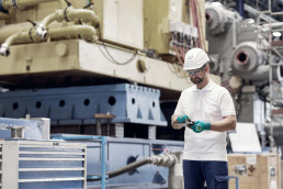 Arbeiter mit uvex Schutzhelm, Schutzbrille und Schutzhandschuhen vor Maschinen in einer Industriehalle.