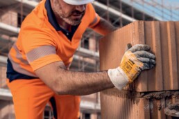 Bauarbeiter mit uvex Schutzhelm und Schutzhandschuhen setzt auf einer Baustelle Ziegel aufeinander.