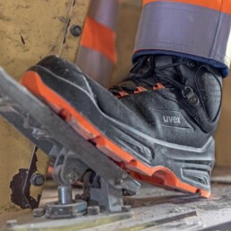 Nahaufnahme-eines-schwarzen-UVEX-Sicherheitsstiefels-mit-orangefarbenen-Akzenten-auf-einem-Industriepedal,-getragen-von-einer-Person-in-Warnschutzkleidung