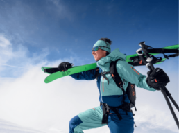 Skifahrer trägt Skiausrüstung über der Schulter, in winterlicher Berglandschaft mit blauem Himmel.