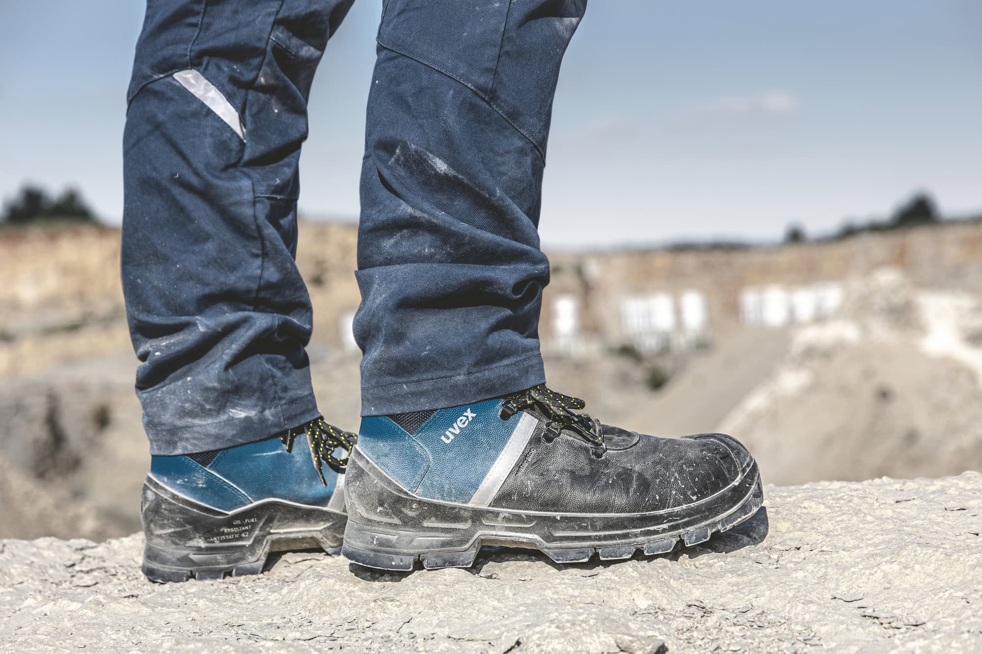Nahaufnahme von uvex 2 construction Sicherheitsschuhen, getragen von einer Person in blauer Arbeitskleidung. Die Schuhe stehen auf einem staubigen Untergrund in einer Baustellenumgebung, im Hintergrund ist eine Steinbruchlandschaft zu erkennen.