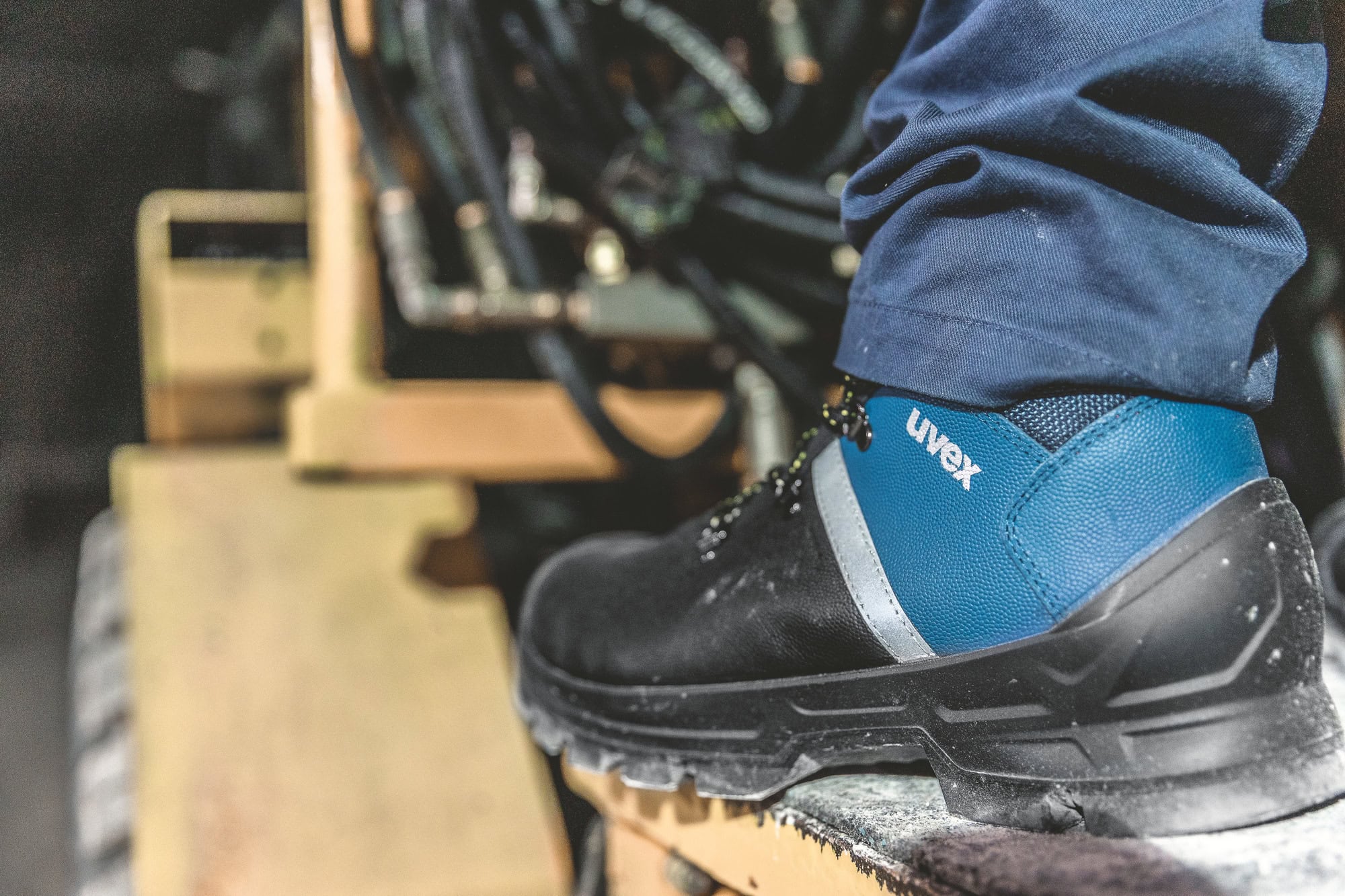 Detailansicht eines uvex 2 construction Sicherheitsschuhs mit blauem und schwarzem Design. Der Schuh steht auf einer Metallplattform, umgeben von hydraulischen Schläuchen und Maschinenkomponenten.