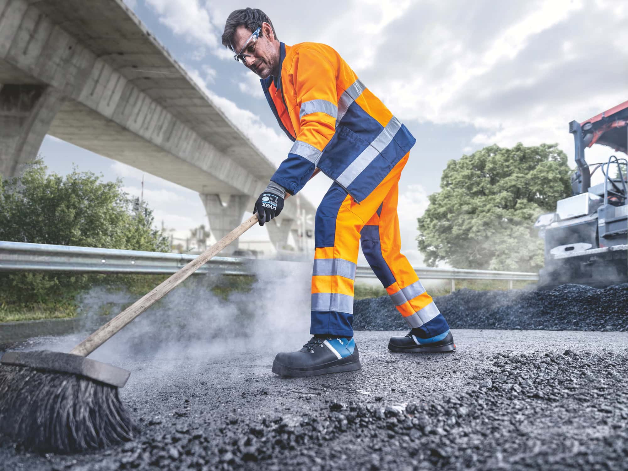 Ein Straßenarbeiter in orange-blauer Schutzkleidung kehrt frisch verlegten Asphalt mit einem Besen. Er trägt uvex 3 asphaltpro Sicherheitsschuhe, die für Arbeiten in heißen und anspruchsvollen Umgebungen geeignet sind. Im Hintergrund sind eine Brücke, grüne Bäume und eine Asphaltiermaschine zu sehen.