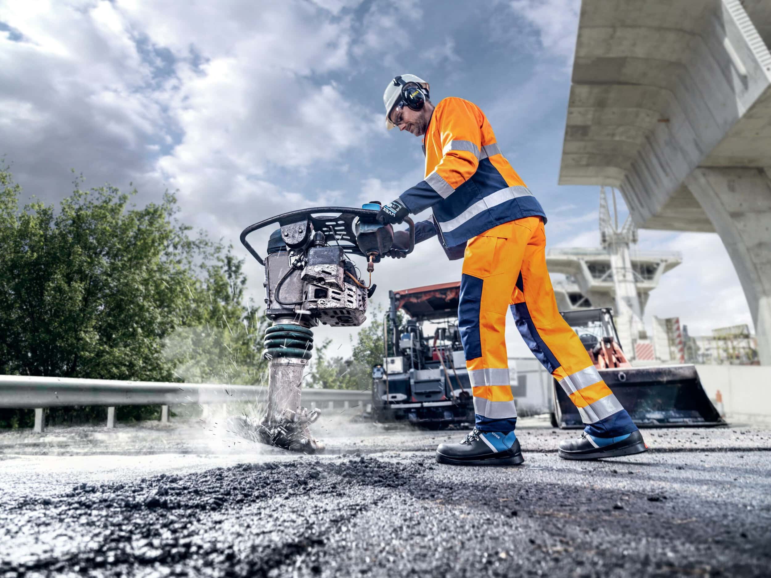 Ein Straßenarbeiter in leuchtender Arbeitskleidung bedient eine Rüttelplatte auf einer asphaltierten Straße, während im Hintergrund Baumaschinen und Brückenelemente sichtbar sind.