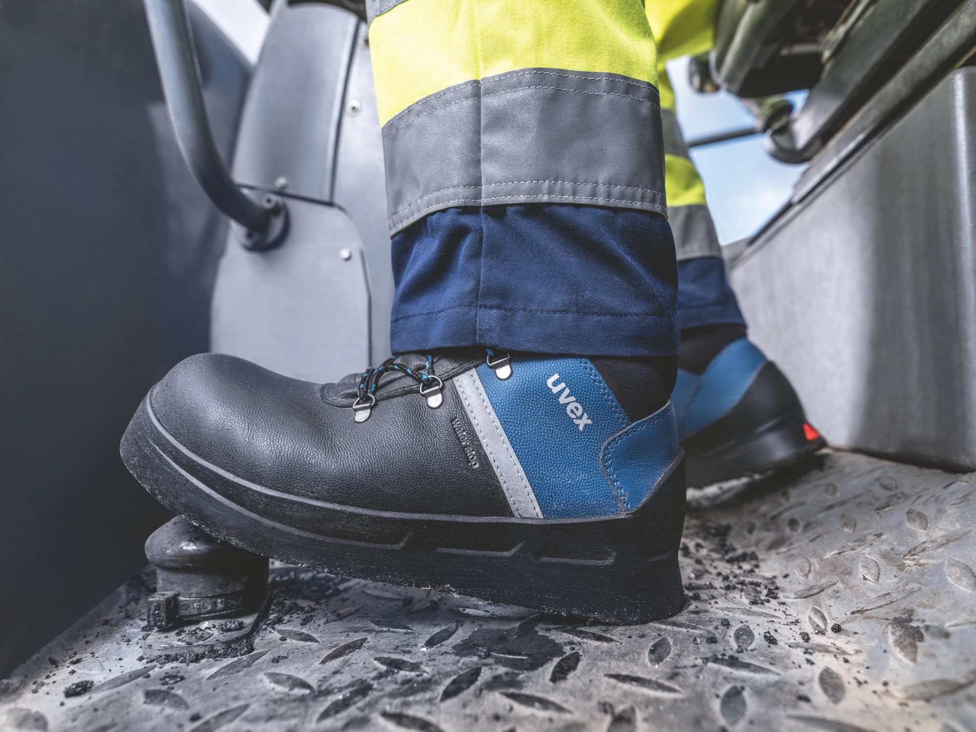 Ein Arbeiter trägt uvex 3 asphaltpro Sicherheitsschuhe mit einer robusten schwarzen Sohle und blauem Obermaterial. Der Schuh steht auf einer rutschfesten Metallplattform, während der Arbeiter gelbe und graue Arbeitskleidung trägt. Die Umgebung deutet auf den Innenraum einer Maschine hin.