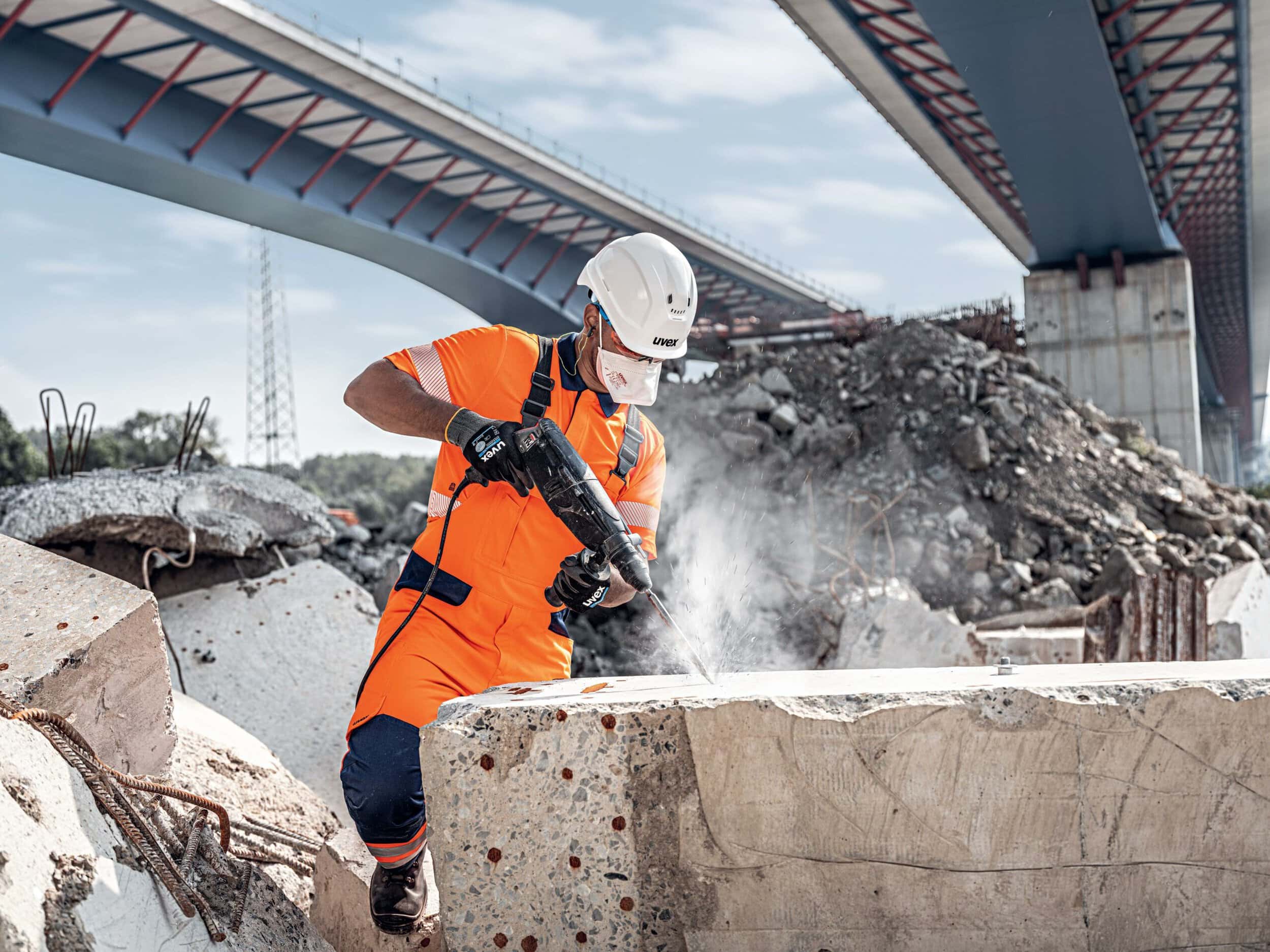 Ein Bauarbeiter in orangefarbener Arbeitskleidung und Schutzhelm verwendet einen Bohrhammer, um Beton unter einer Brücke zu bearbeiten.
