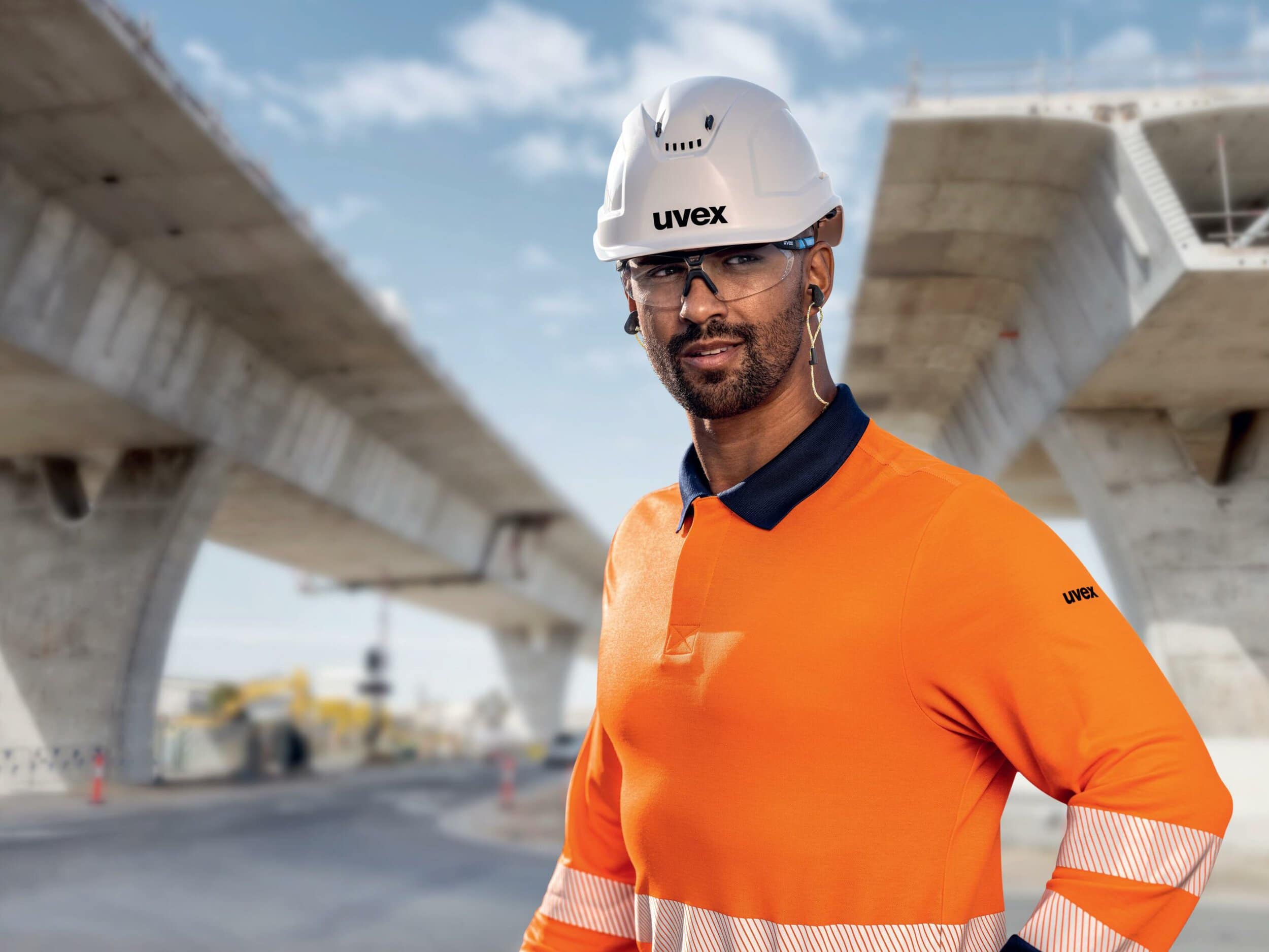 Ein Bauarbeiter in orangefarbener Arbeitskleidung und Schutzhelm steht unter einer großen Brücke, die sich über eine Baustelle erstreckt.