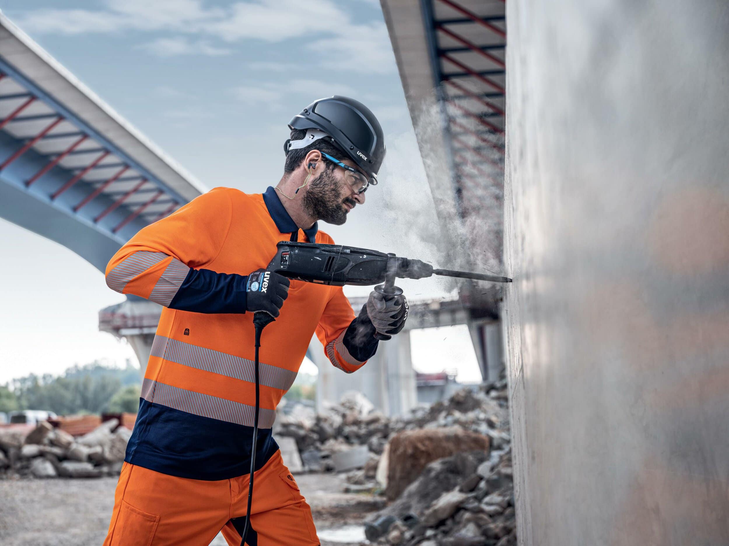 Ein Bauarbeiter in orangefarbener Schutzkleidung und Helm verwendet einen Bohrhammer, um eine Betonwand zu bearbeiten, mit Brückenstrukturen und Trümmern im Hintergrund.