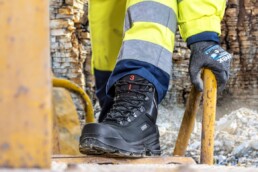 Ein Arbeiter trägt schwarze uvex quatro 3 Sicherheitsschuhe mit einem robusten Design. Die Schuhe sind mit einer rutschfesten Sohle ausgestattet und werden in einer Baustellenumgebung getragen.
