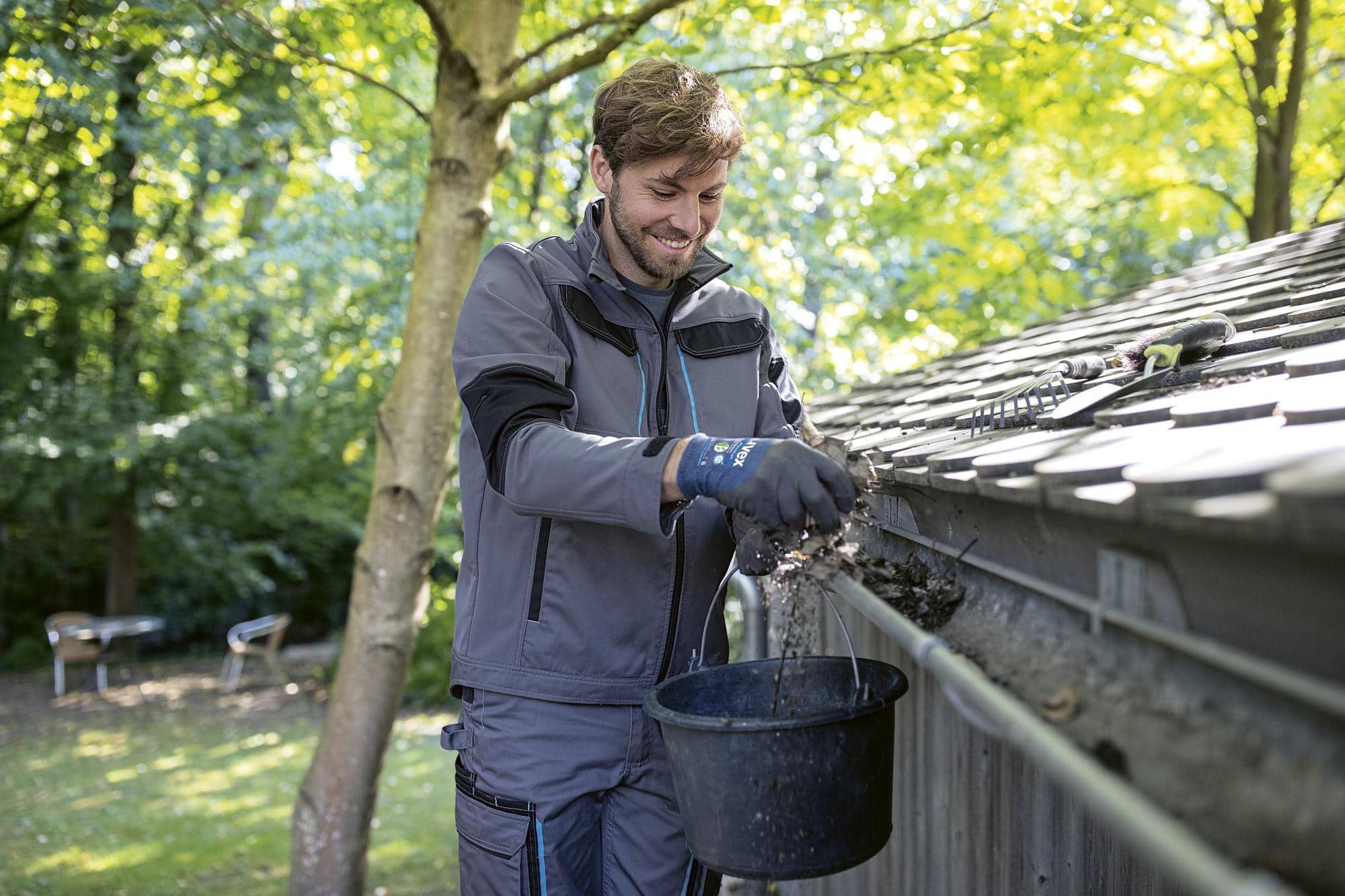 Mann in Arbeitskleidung reinigt Dachrinne mit Handschuhen und Eimer im Garten.