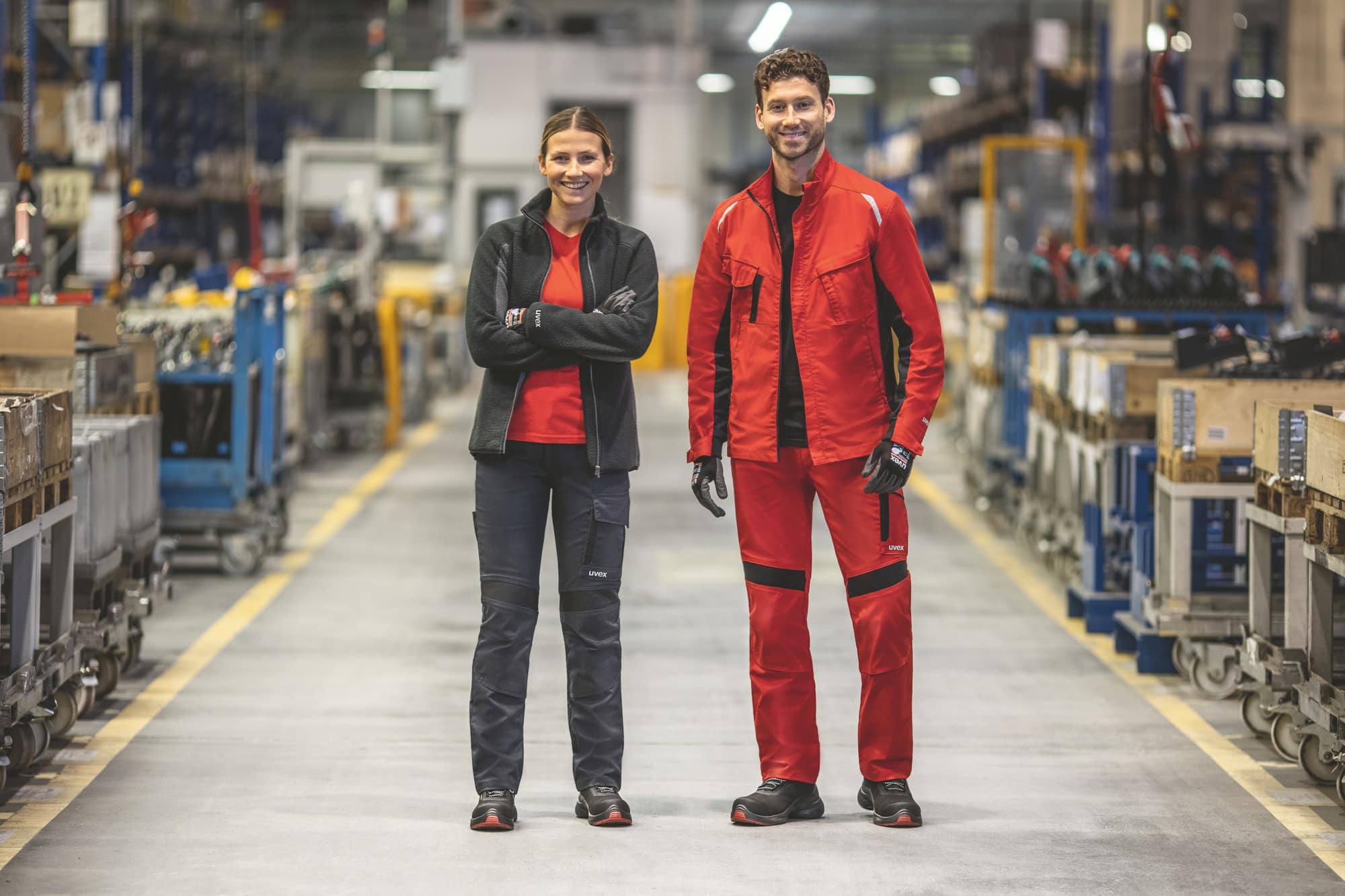 Zwei Personen in roter Arbeitskleidung stehen in einer Fabrikhalle mit Paletten.