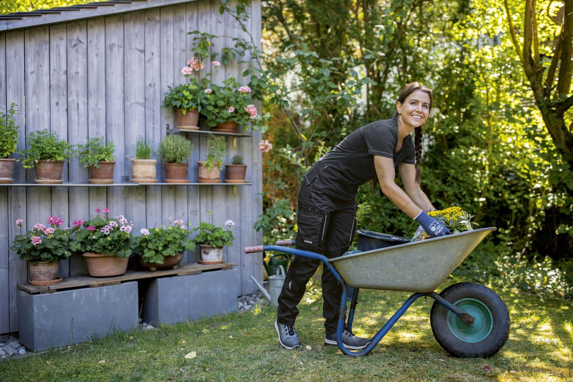 Frau in Arbeitskleidung schiebt Schubkarre mit Gartenabfällen vor Holzwand mit Pflanzen.