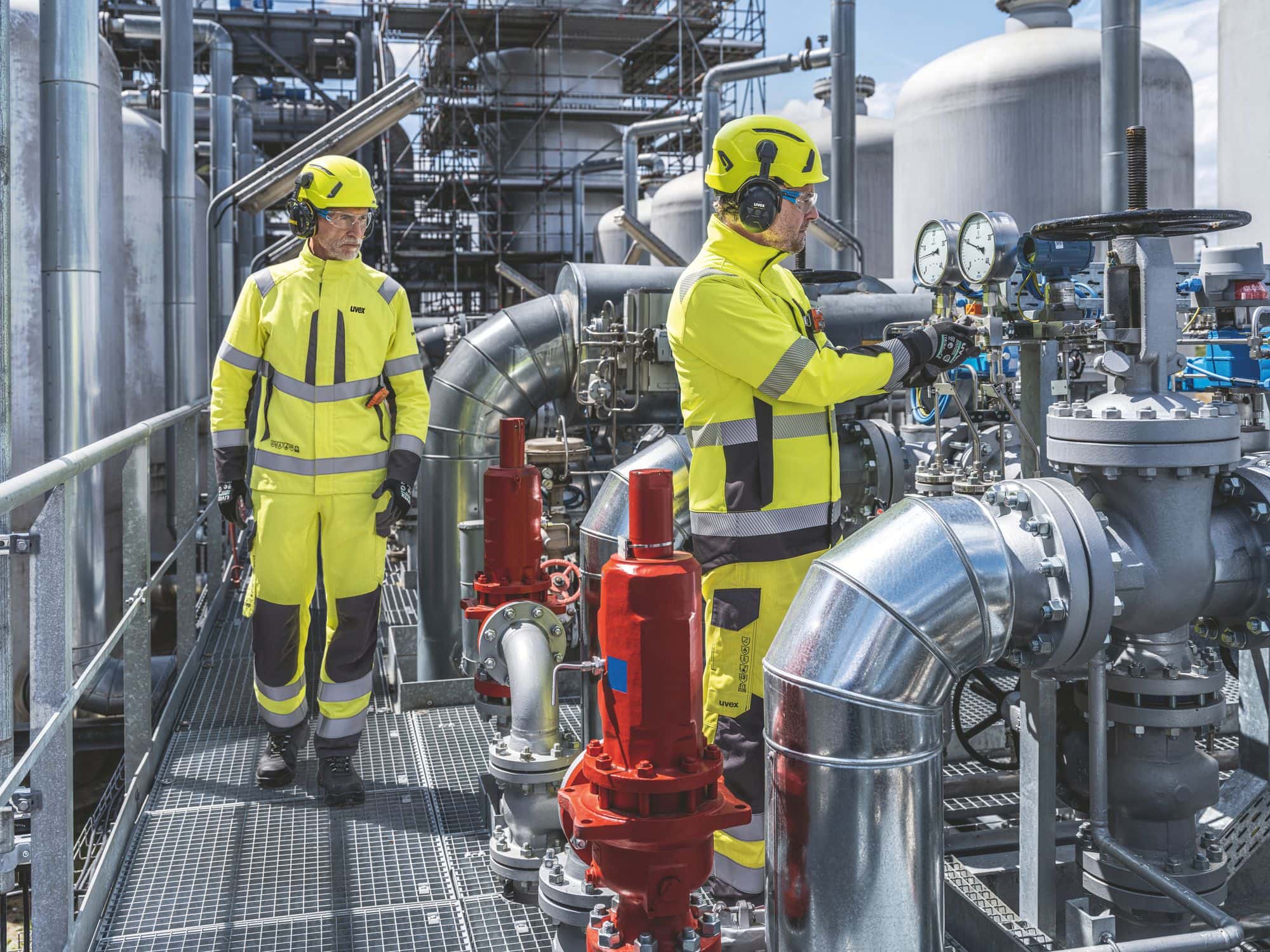 Zwei Arbeiter in gelber Schutzkleidung mit Helmen in einer Industrieanlage mit Rohren.