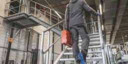 Ein Arbeiter in grauer Arbeitskleidung und Handschuhen steigt mit einem orangefarbenen Werkzeugkoffer eine Metalltreppe in einer Industriehalle hinauf.