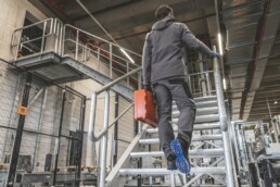 Ein Arbeiter in grauer Arbeitskleidung und Handschuhen steigt mit einem orangefarbenen Werkzeugkoffer eine Metalltreppe in einer Industriehalle hinauf.