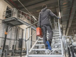 Ein Arbeiter in grauer Arbeitskleidung und Handschuhen steigt mit einem orangefarbenen Werkzeugkoffer eine Metalltreppe in einer Industriehalle hinauf.