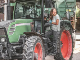 Eine Frau mit langen Haaren und blauen Handschuhen steigt in Arbeitskleidung auf einen großen grünen Traktor mit roten Felgen, der auf einem Bauernhof steht.
