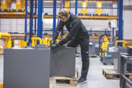Ein Mann in schwarzer Arbeitskleidung befestigt in einer Lagerhalle mit Handschuhen einen Hebegurt.