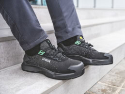 Eine Person sitzt auf hellen Steinstufen und trägt moderne, schwarz-grüne uvex-Sicherheitsschuhe mit gestricktem Obermaterial und gepolstertem Schaft.