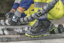 Ein Bauarbeiter trägt schwarze uvex-Sicherheitsschuhe mit neongrüner Sohle und uvex-Schutzhandschuhe, während er mit großen, staubigen Kabeln oder Schläuchen auf einer Baustelle hantiert.
